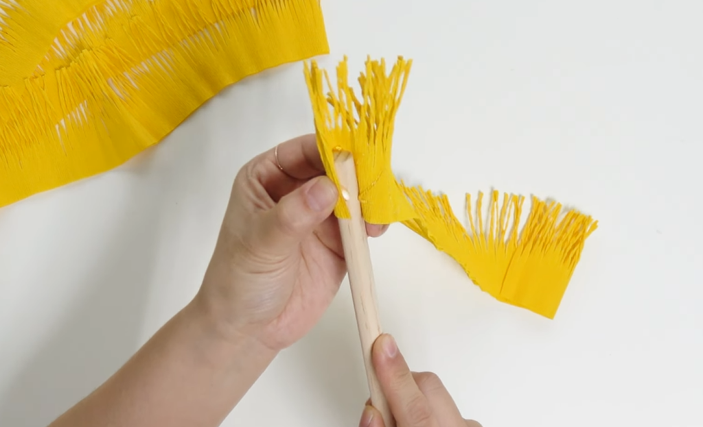 Wrapping yellow crepe paper fringe around a wooden stick to form a giant daisy center