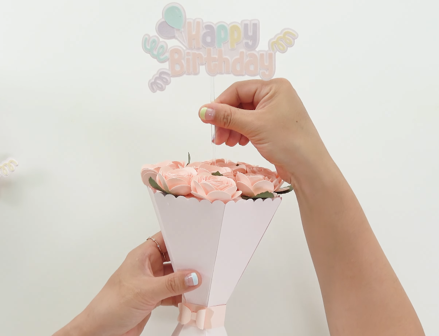Placing a colorful “Happy Birthday” topper into the finished pink paper flower bouquet.