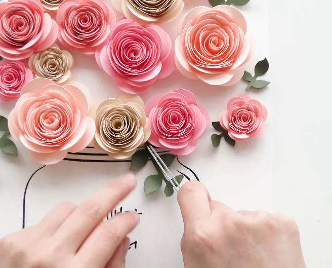 Arranging green paper leaves between pink rolled paper roses on a floral wall art frame