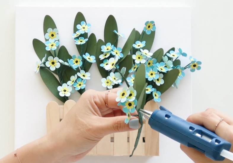 Applying hot glue to forget-me-not flower stems before attaching them to the canvas