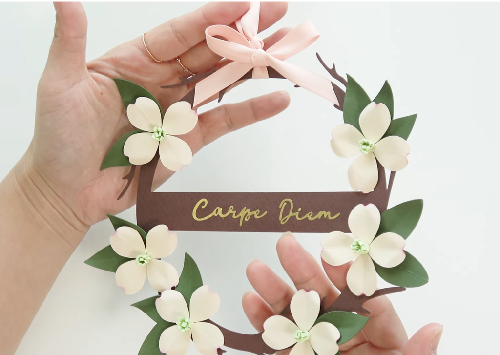 Finished DIY dogwood paper flower wreath with ivory flowers, green leaves, a pink ribbon, and gold Carpe Diem lettering