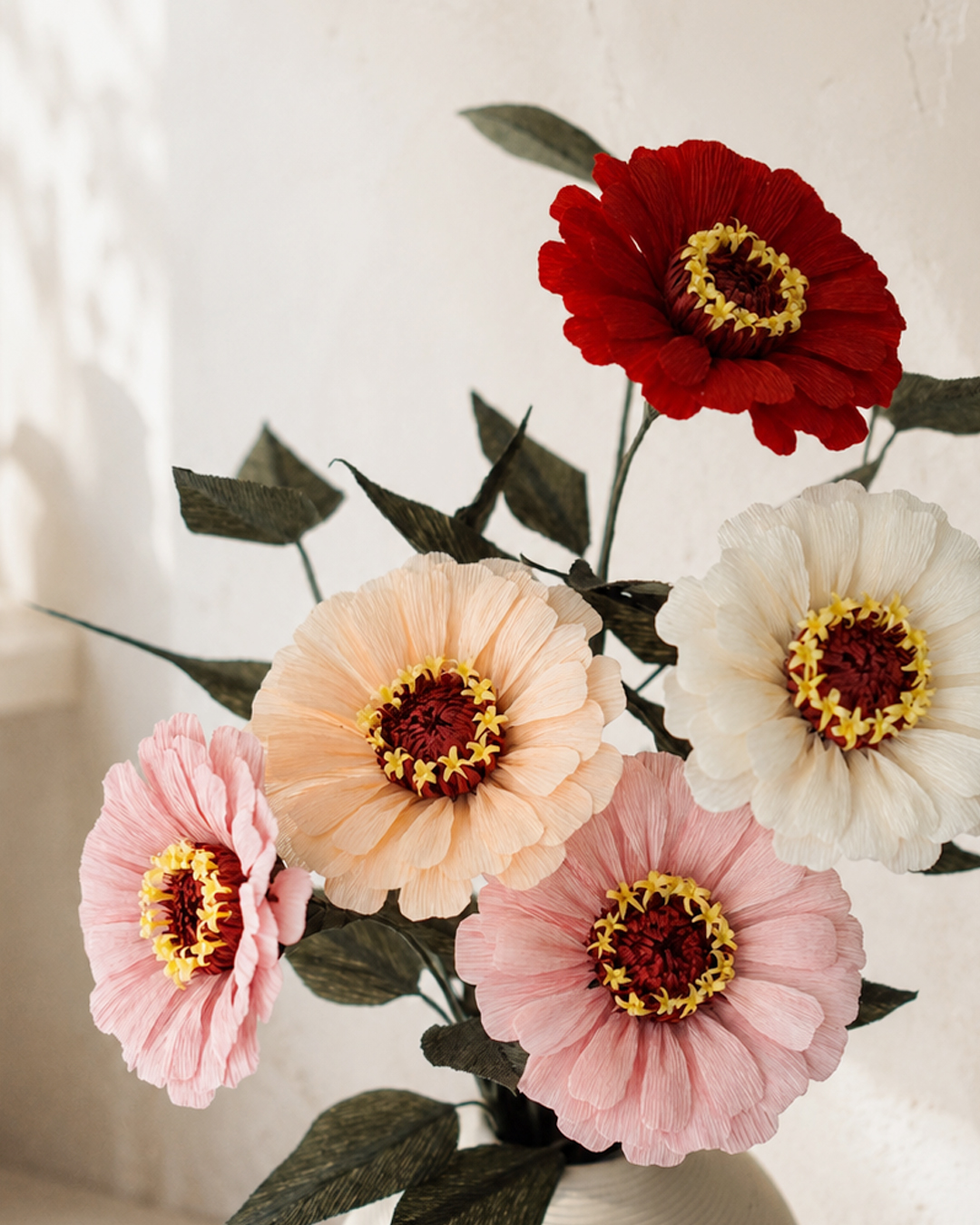 Handmade crepe paper zinnia flowers in red, pink, peach, and white arranged in a vase with dark green leaves.