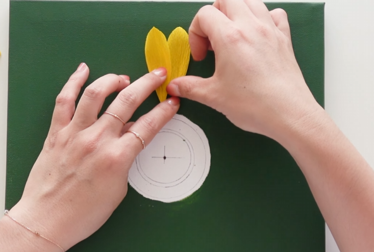 Attaching the first layer of yellow crepe paper sunflower petals to the green canvas
